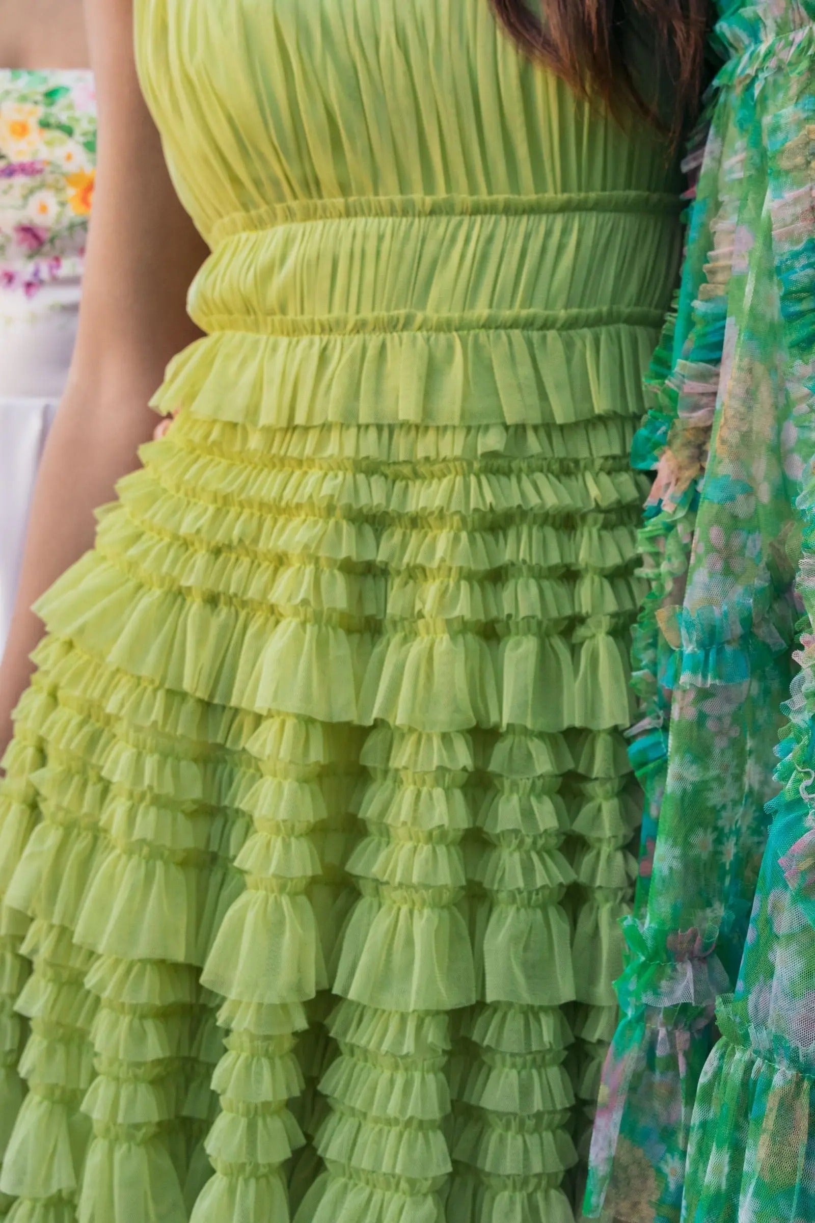 Close-up of a person wearing the light green Mac Duggal 8292 tea-length party dress, featuring mesh fabric and ruffle details, standing next to another person in a multicolored floral outfit.
